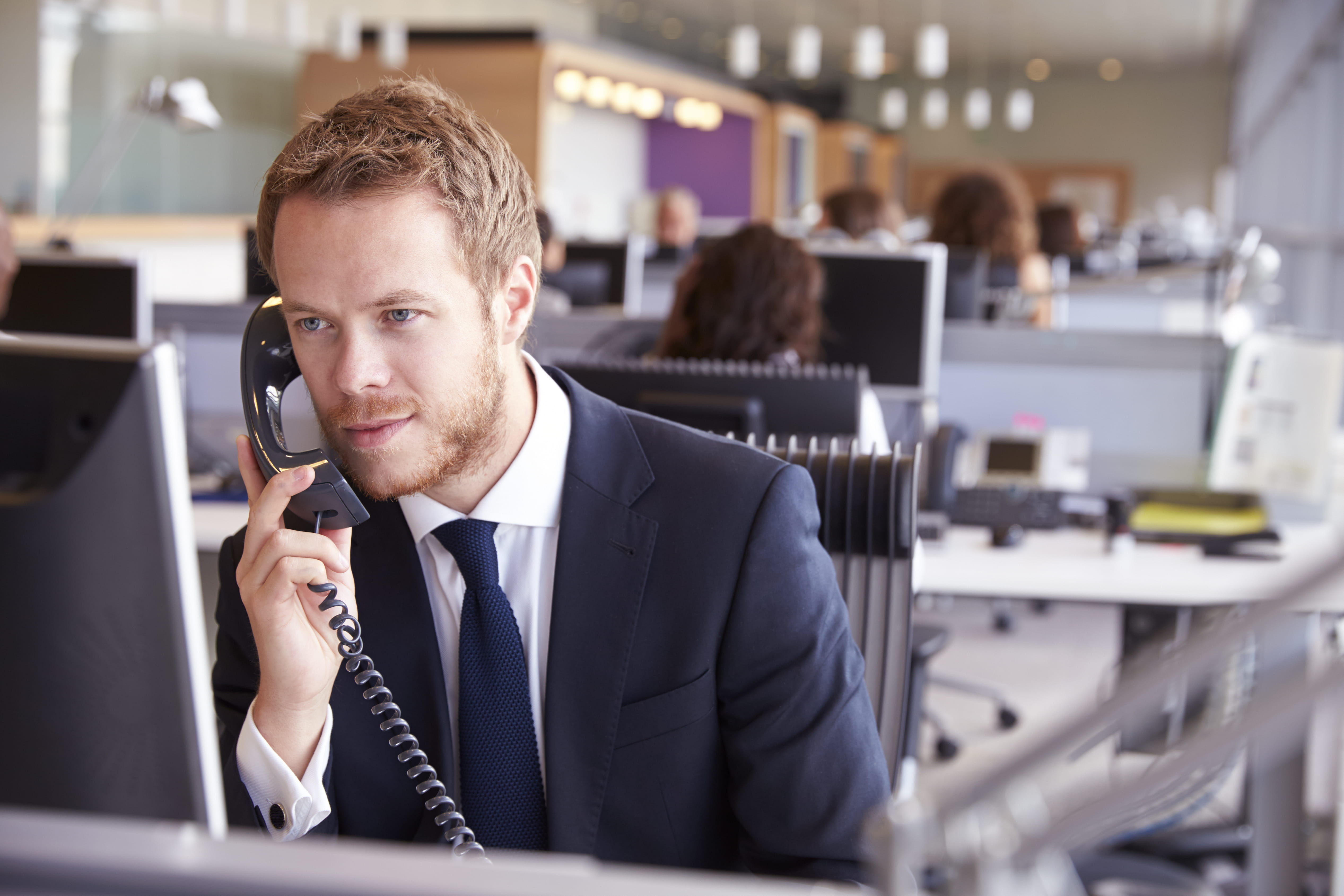 A young man wearing a suit answers questions on the telephone sitting in an office.