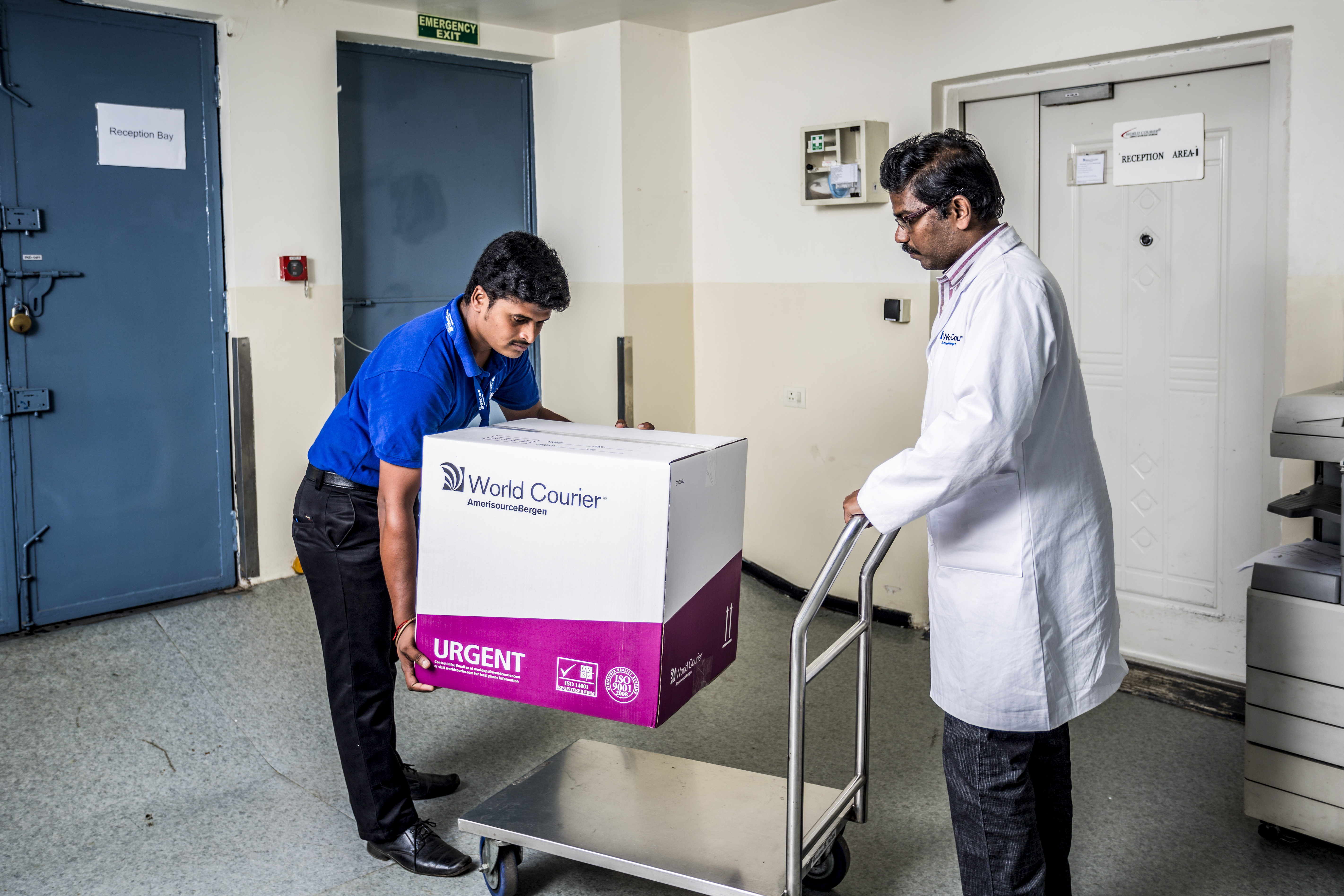 A World Courier driver loads a large box onto a cart pushed by a man in a lab coat.